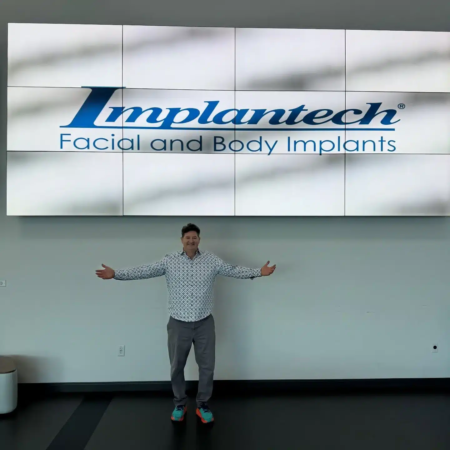 Dr. Robert J. Troell standing with his arms outstretched beneath the Implantech Facial and Body Implants sign at the Implantech Masters Educational Series, November 2025, where he serves as Distinguished Faculty